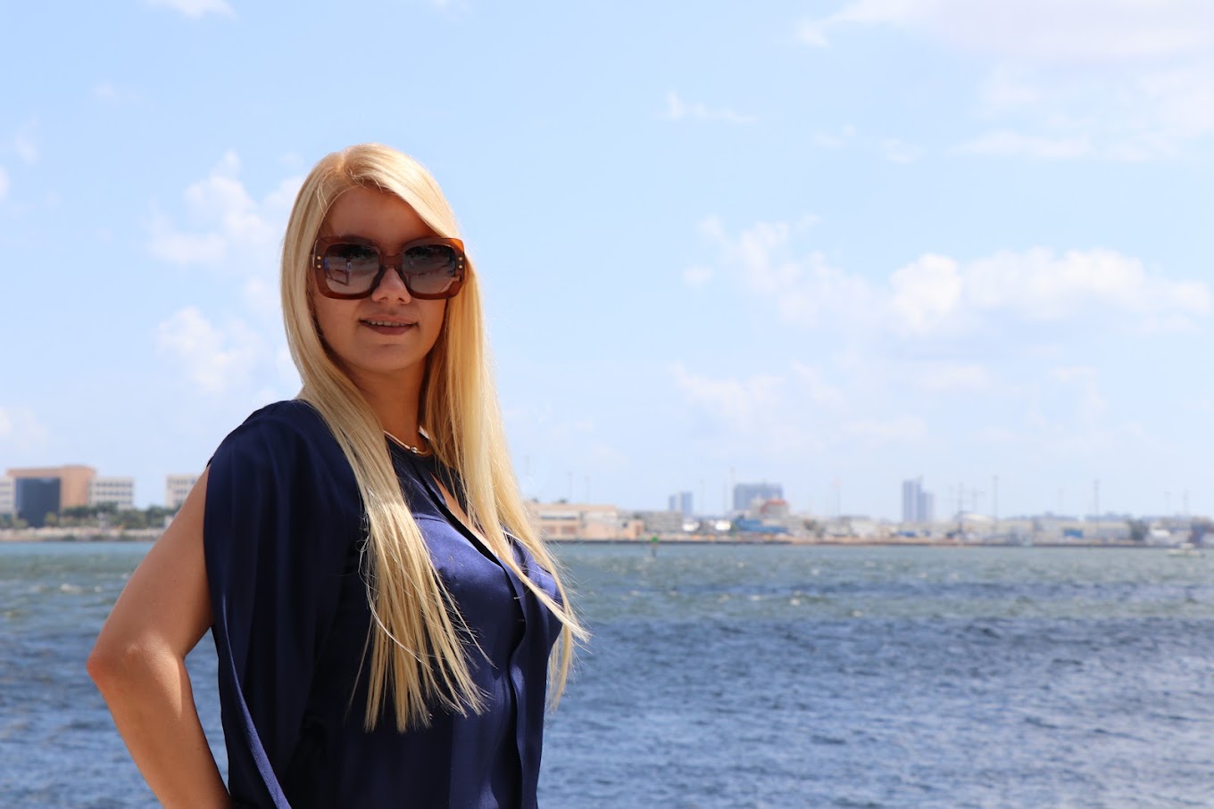 Una persona de cabello largo, con blusa azul marino y lentes de sol, al aire libre junto al agua y con la costa de la ciudad al fondo, durante una sesión fotográfica corporativa de Safeguard and Associates.
